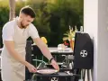 Mężczyzna w fartuchu przygotowuje mięso na grillu na balkonie.