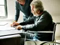 Starszy mężczyzna na wózku inwalidzkim, z pomocą opiekunki, podpisuje dokumenty. To może być ważne pełnomocnictwo od osoby z demencją.