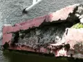 Zniszczony balkon w bloku z wielkiej płyty, z odpadającą czerwoną farbą i widoczną cegłą.