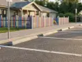 Pusta nawierzchnia asfaltowa z wyznaczonymi białymi liniami i betonowymi odbojnikami. W tle kolorowy płotek i budynek. Widać, że szerokość miejsca parkingowego jest standardowa.
