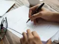 Dłoń pisze długopisem na dokumencie. Obok leżą okulary. To może być pełnomocnictwo notarialne osoby chorej, przygotowywane do podpisania.