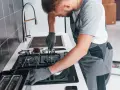 Fachowiec w rękawiczkach montuje podłączenie kuchenki gazowej w bloku. Narzędzia leżą obok.