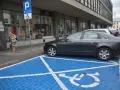 Niebieskie miejsce parkingowe dla niepełnosprawnych z białym symbolem wózka inwalidzkiego. Obok stoi ciemny samochód.