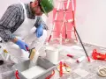 Malowanie ścian w bloku. Remont mieszkania w jakich godzinach? Malarz w białym kombinezonie zanurza pędzel w farbie.