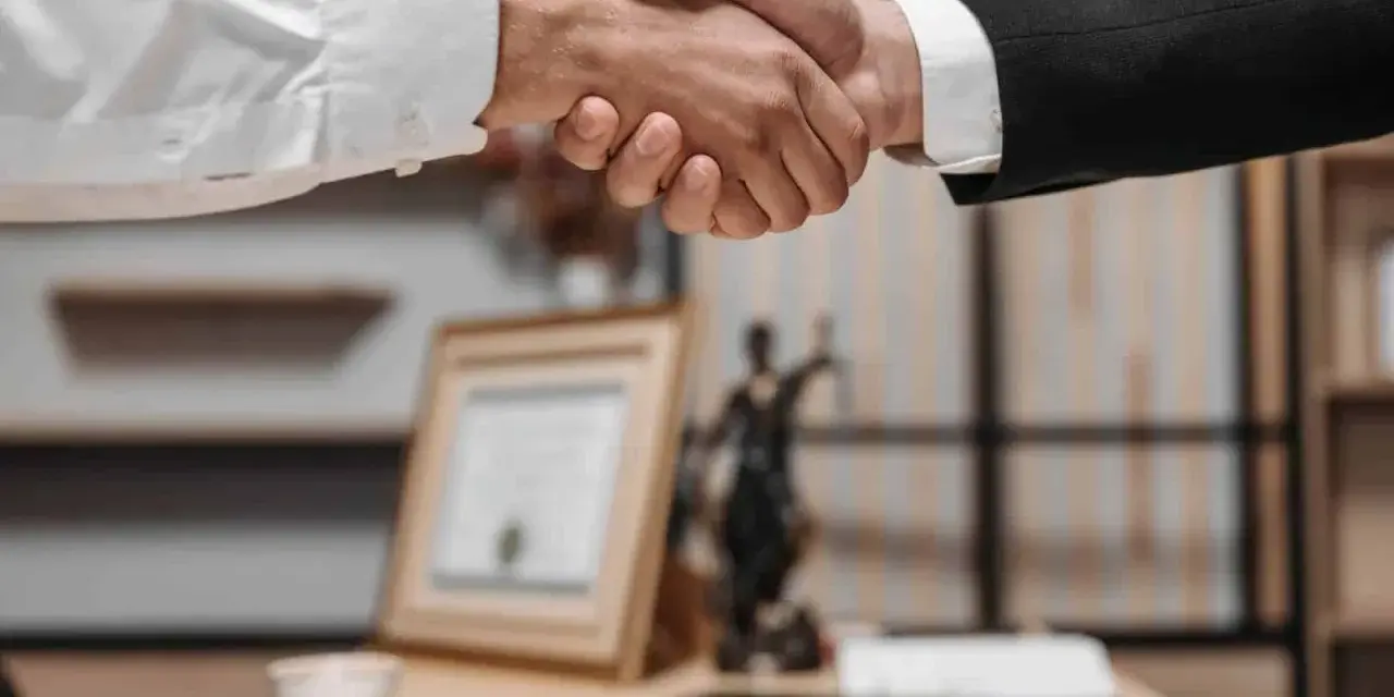 Uścisk dłoni między dwoma mężczyznami w garniturach. W tle widać statuetkę Temidy i dokumenty, co sugeruje spotkanie z radcą prawnym nieruchomości.