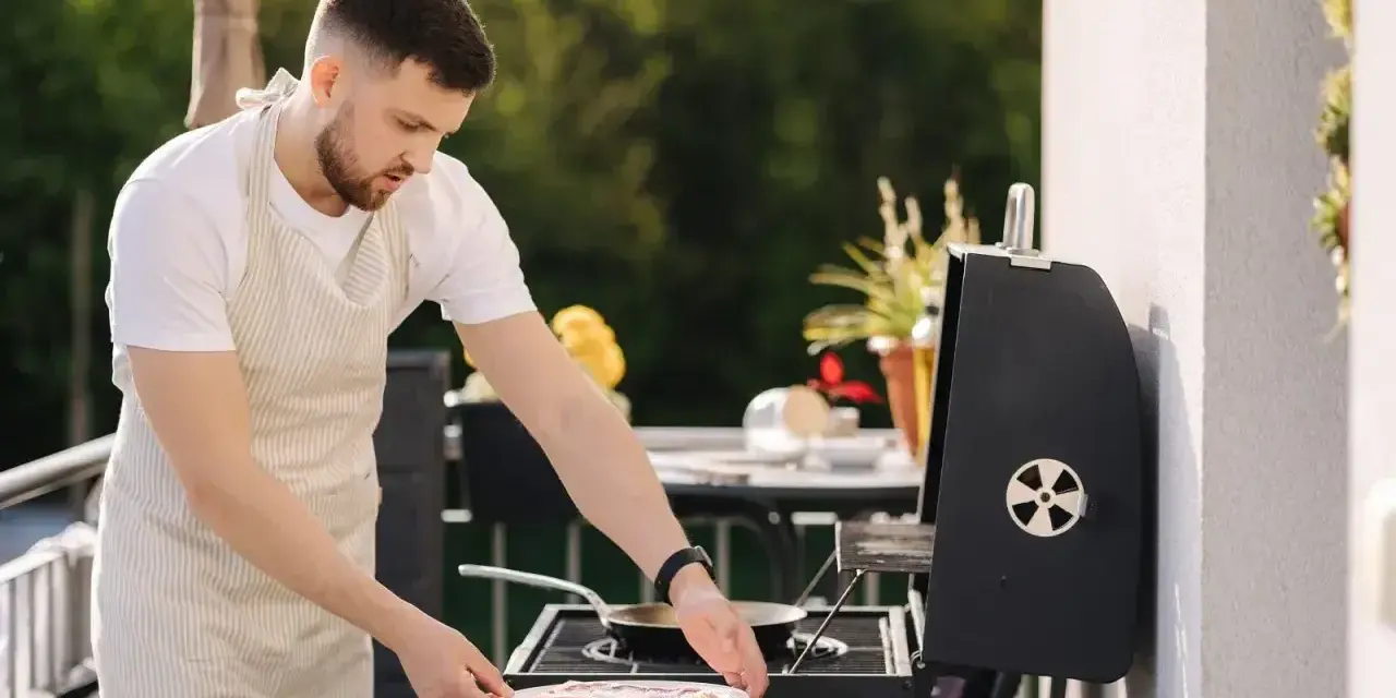 Mężczyzna w fartuchu przygotowuje mięso na grillu na balkonie.