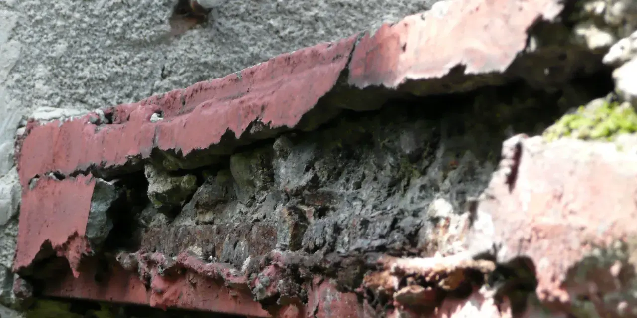 Zniszczony balkon w bloku z wielkiej płyty, z odpadającą czerwoną farbą i widoczną cegłą.