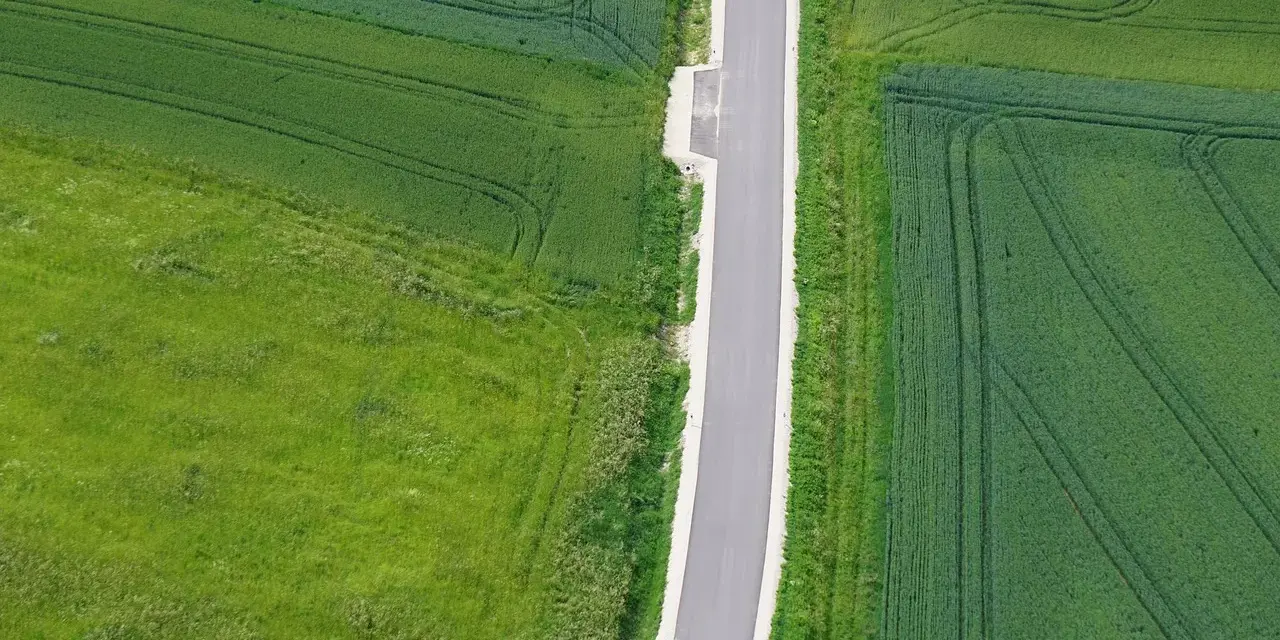 Wąska droga dojazdowa do działki, otoczona polami zielonych zbóż i traw.