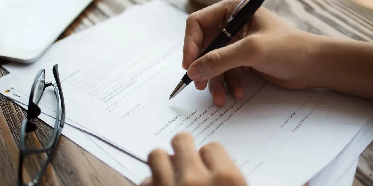 Dłoń pisze długopisem na dokumencie. Obok leżą okulary. To może być pełnomocnictwo notarialne osoby chorej, przygotowywane do podpisania.