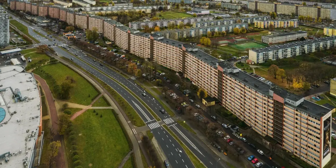 Najdłuższy blok w Polsce, falujący budynek mieszkalny z pomarańczową elewacją, otoczony zielenią i drogą z samochodami.