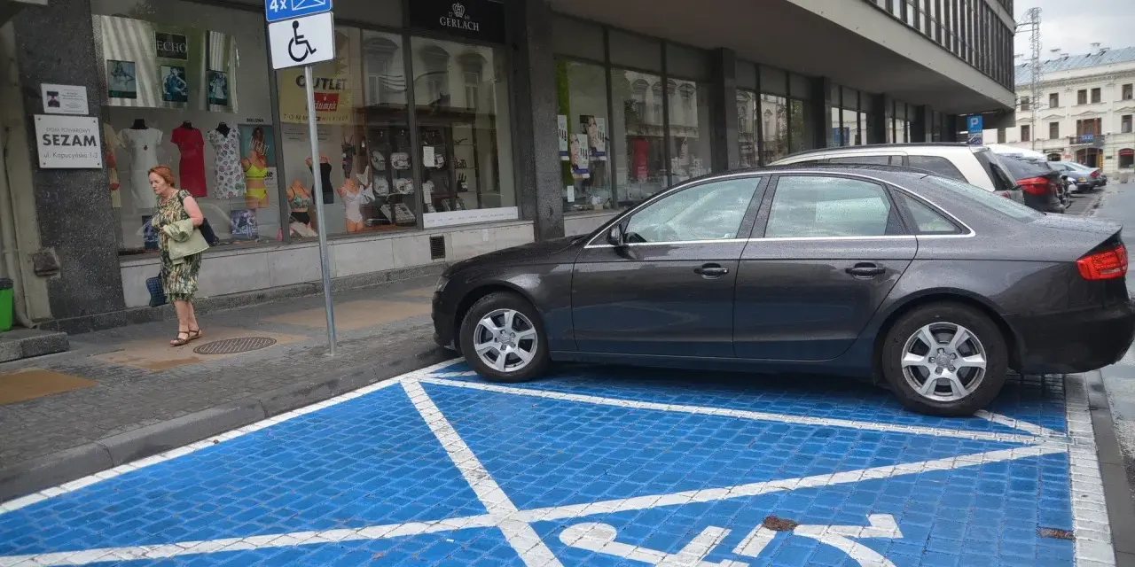 Niebieskie miejsce parkingowe dla niepełnosprawnych z białym symbolem wózka inwalidzkiego. Obok stoi ciemny samochód.