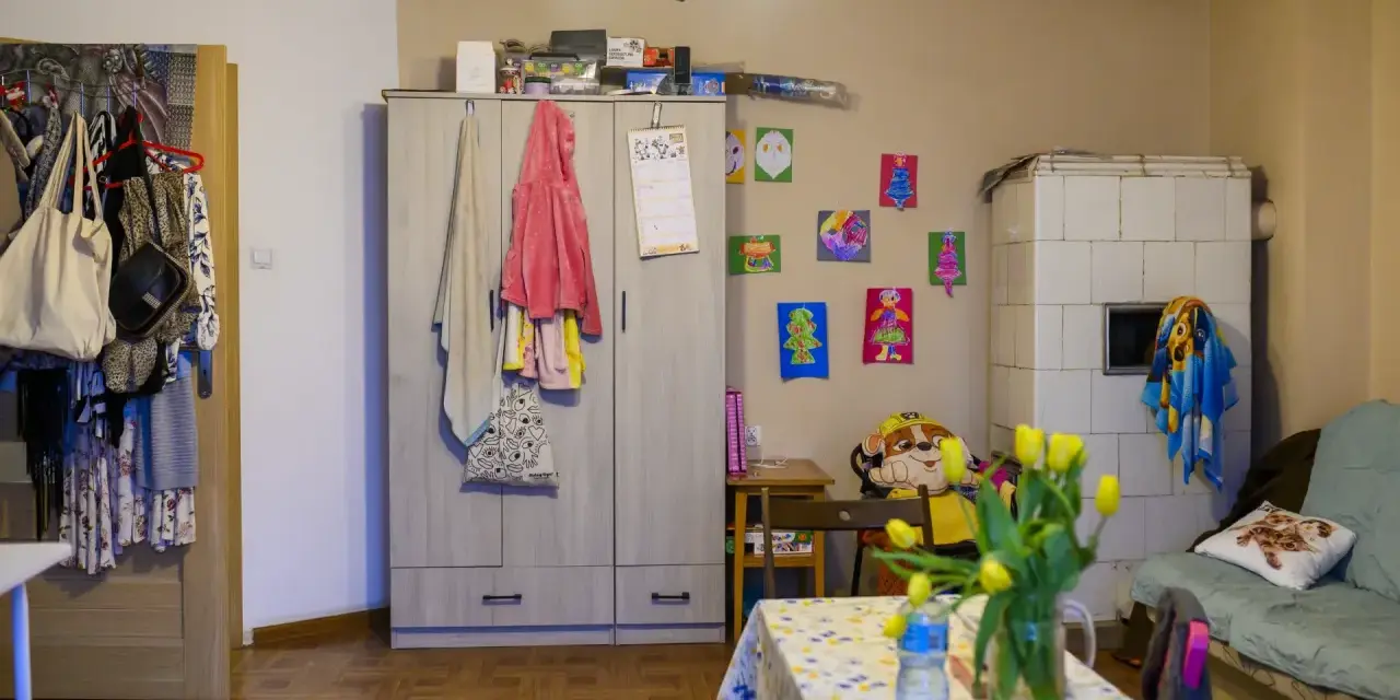 Przytulne mieszkanie dla samotnej matki. Szafa, piec kaflowy, kanapa, stolik z tulipanami i dziecięce rysunki na ścianie tworzą domową atmosferę.
