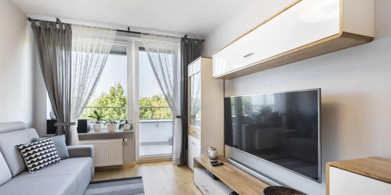 Przytulny, bardzo mały salon w bloku z szarą kanapą, telewizorem i oknem balkonowym.