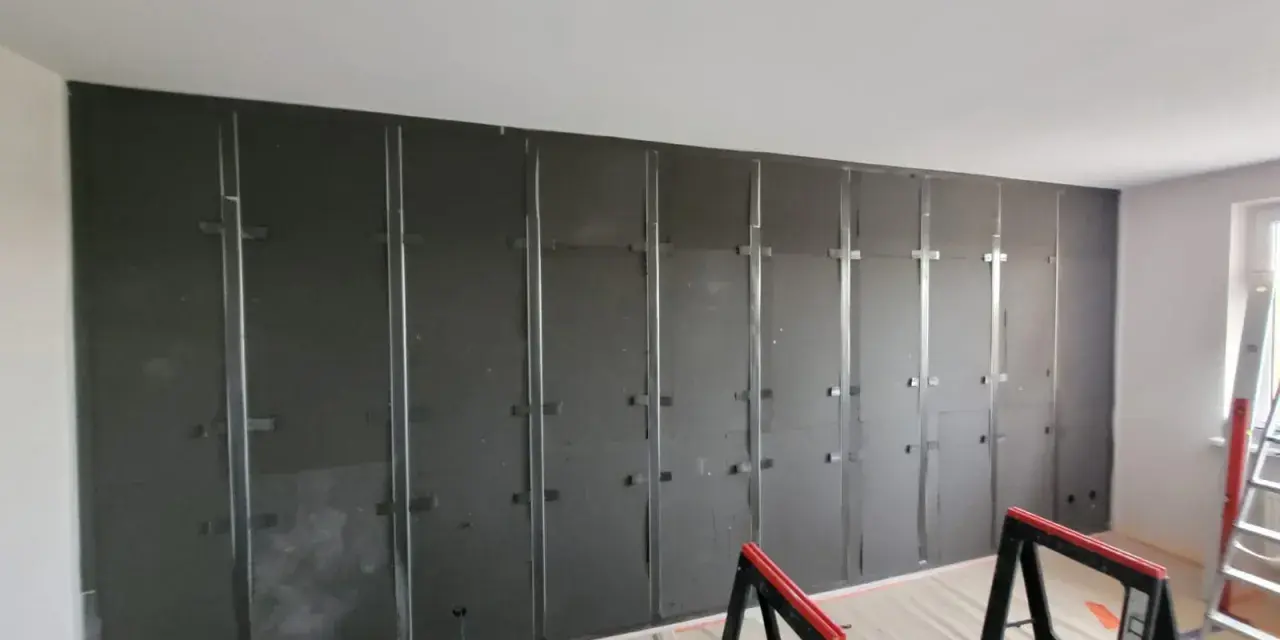 Montaż paneli wygłuszających ścianę od sąsiada. Metalowy stelaż i ciemne płyty tworzą barierę akustyczną.