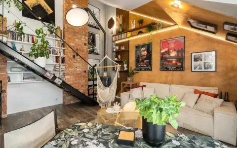 Przytulne mieszkanie w stylu loft z ceglaną ścianą, metalowymi schodami, wiszącym fotelem i plakatami filmowymi.