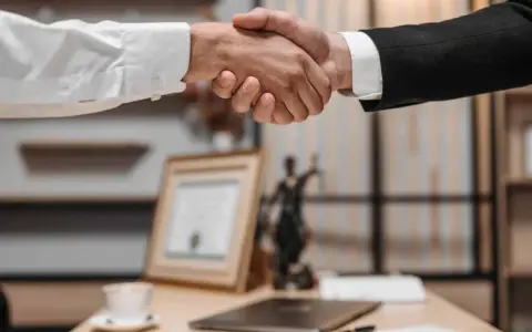 Uścisk dłoni między dwoma mężczyznami w garniturach. W tle widać statuetkę Temidy i dokumenty, co sugeruje spotkanie z radcą prawnym nieruchomości.