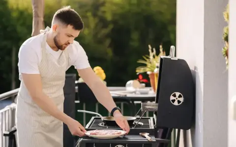 Mężczyzna w fartuchu przygotowuje mięso na grillu na balkonie.