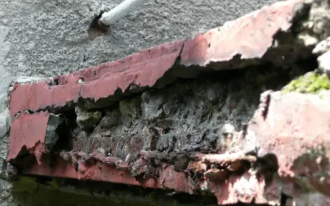 Zniszczony balkon w bloku z wielkiej płyty, z odpadającą czerwoną farbą i widoczną cegłą.