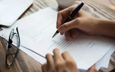 Dłoń pisze długopisem na dokumencie. Obok leżą okulary. To może być pełnomocnictwo notarialne osoby chorej, przygotowywane do podpisania.