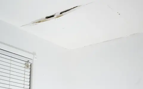 Pęknięcie na suficie w bloku, widoczne fragmenty tynku i izolacji. Obok okno z żaluzjami.