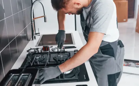 Fachowiec w rękawiczkach montuje podłączenie kuchenki gazowej w bloku. Narzędzia leżą obok.