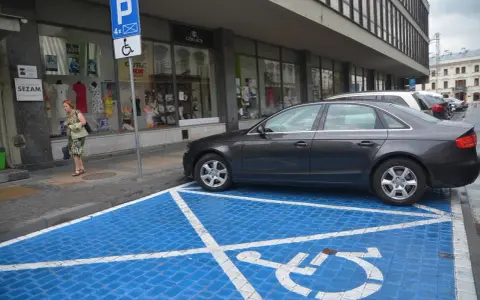 Niebieskie miejsce parkingowe dla niepełnosprawnych z białym symbolem wózka inwalidzkiego. Obok stoi ciemny samochód.