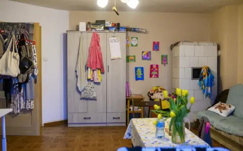 Przytulne mieszkanie dla samotnej matki. Szafa, piec kaflowy, kanapa, stolik z tulipanami i dziecięce rysunki na ścianie tworzą domową atmosferę.