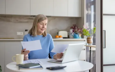Kobieta analizuje dokumenty, porównując je z informacjami na laptopie. Może to dotyczyć spraw związanych z zameldowaniem a zasiedzeniem.