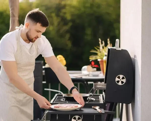 Mężczyzna w fartuchu przygotowuje mięso na grillu na balkonie.