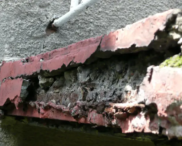 Zniszczony balkon w bloku z wielkiej płyty, z odpadającą czerwoną farbą i widoczną cegłą.