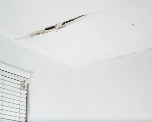 Pęknięcie na suficie w bloku, widoczne fragmenty tynku i izolacji. Obok okno z żaluzjami.