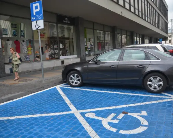 Niebieskie miejsce parkingowe dla niepełnosprawnych z białym symbolem wózka inwalidzkiego. Obok stoi ciemny samochód.