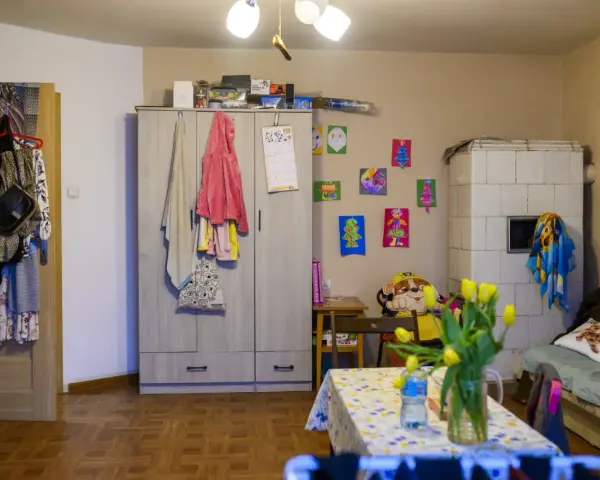 Przytulne mieszkanie dla samotnej matki. Szafa, piec kaflowy, kanapa, stolik z tulipanami i dziecięce rysunki na ścianie tworzą domową atmosferę.