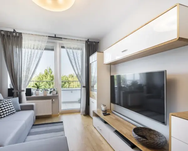 Przytulny, bardzo mały salon w bloku z szarą kanapą, telewizorem i oknem balkonowym.