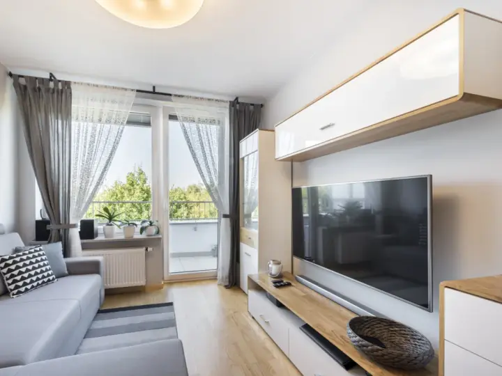 Przytulny, bardzo mały salon w bloku z szarą kanapą, telewizorem i oknem balkonowym.