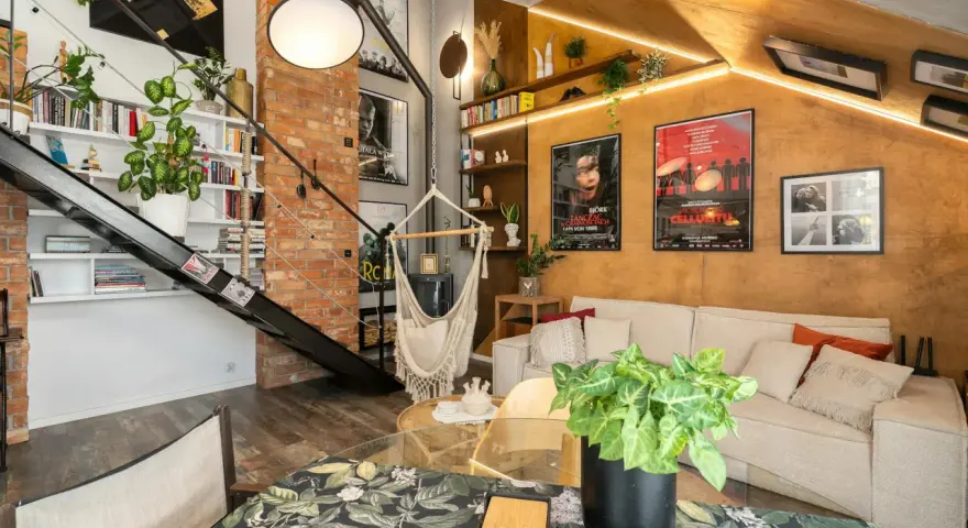 Przytulne mieszkanie w stylu loft z ceglaną ścianą, metalowymi schodami, wiszącym fotelem i plakatami filmowymi.