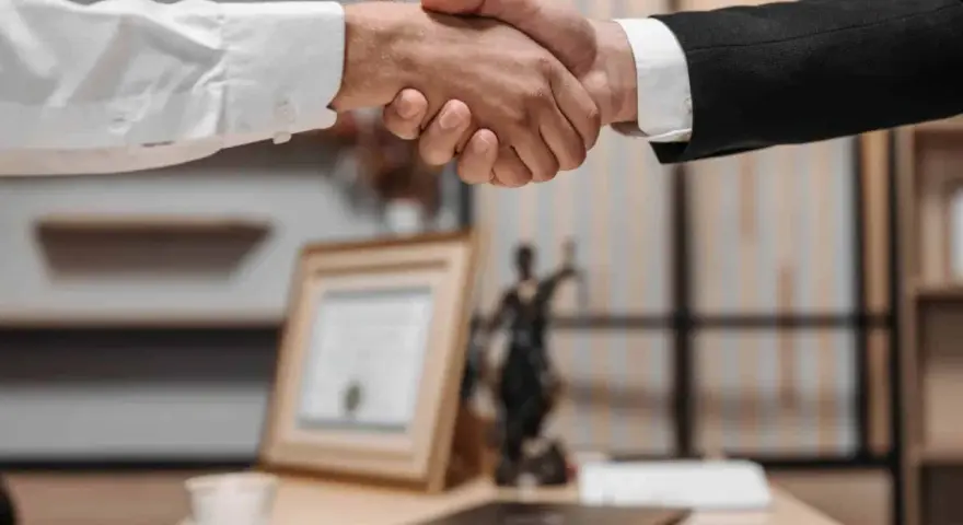 Uścisk dłoni między dwoma mężczyznami w garniturach. W tle widać statuetkę Temidy i dokumenty, co sugeruje spotkanie z radcą prawnym nieruchomości.