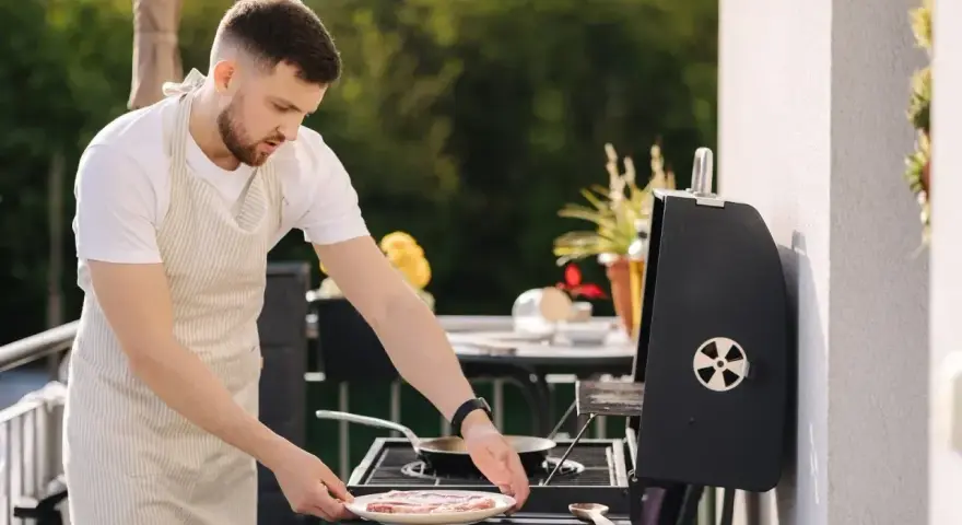 Mężczyzna w fartuchu przygotowuje mięso na grillu na balkonie.