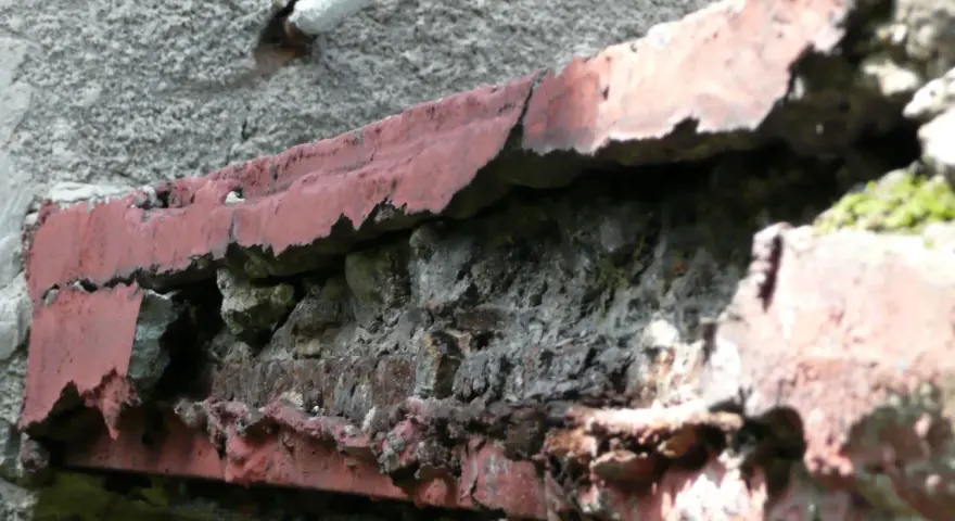 Zniszczony balkon w bloku z wielkiej płyty, z odpadającą czerwoną farbą i widoczną cegłą.