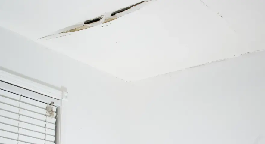 Pęknięcie na suficie w bloku, widoczne fragmenty tynku i izolacji. Obok okno z żaluzjami.