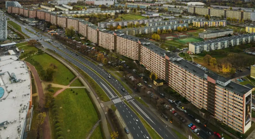 Najdłuższy blok w Polsce, falujący budynek mieszkalny z pomarańczową elewacją, otoczony zielenią i drogą z samochodami.