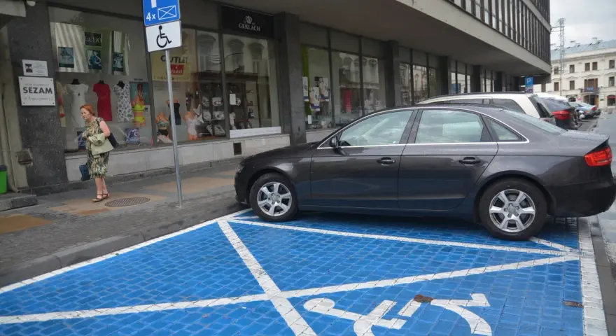 Niebieskie miejsce parkingowe dla niepełnosprawnych z białym symbolem wózka inwalidzkiego. Obok stoi ciemny samochód.