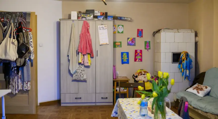 Przytulne mieszkanie dla samotnej matki. Szafa, piec kaflowy, kanapa, stolik z tulipanami i dziecięce rysunki na ścianie tworzą domową atmosferę.