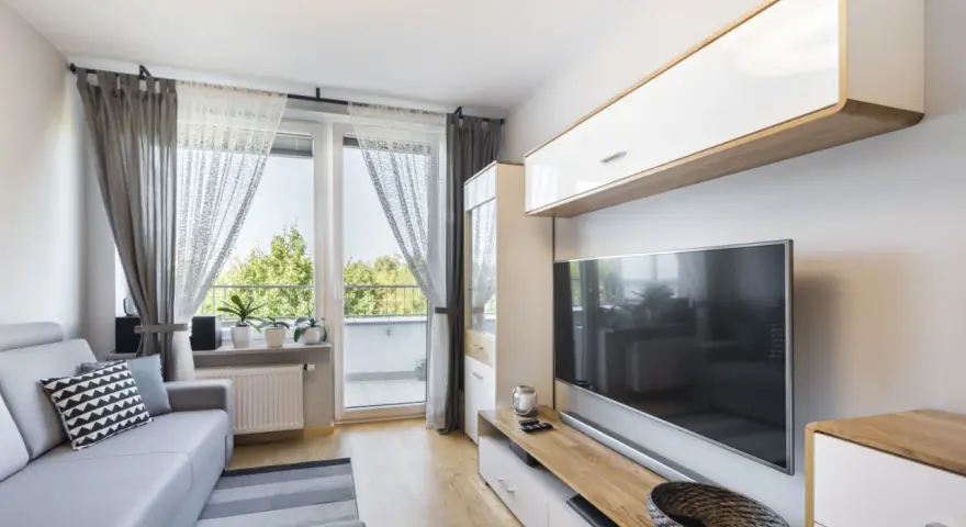 Przytulny, bardzo mały salon w bloku z szarą kanapą, telewizorem i oknem balkonowym.