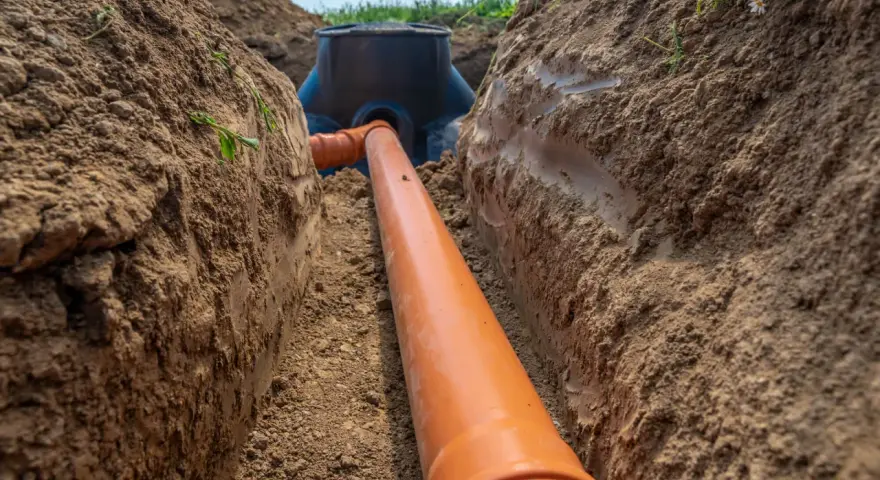 Pomarańczowa rura kanalizacyjna prowadzi do studzienki rewizyjnej sąsiada, zakopana w wykopie.
