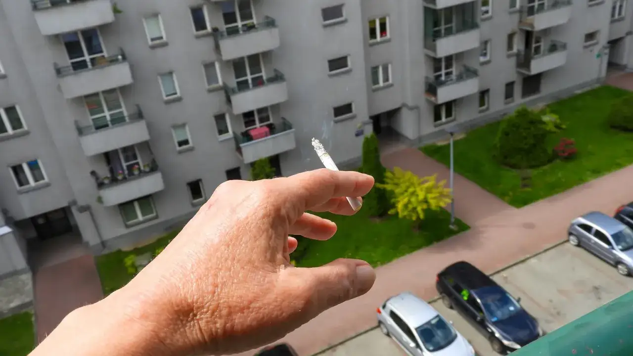 Dłoń trzymająca zapalonego papierosa na tle bloku mieszkalnego. Czy można palić papierosy na balkonie? Dym unosi się w powietrzu.