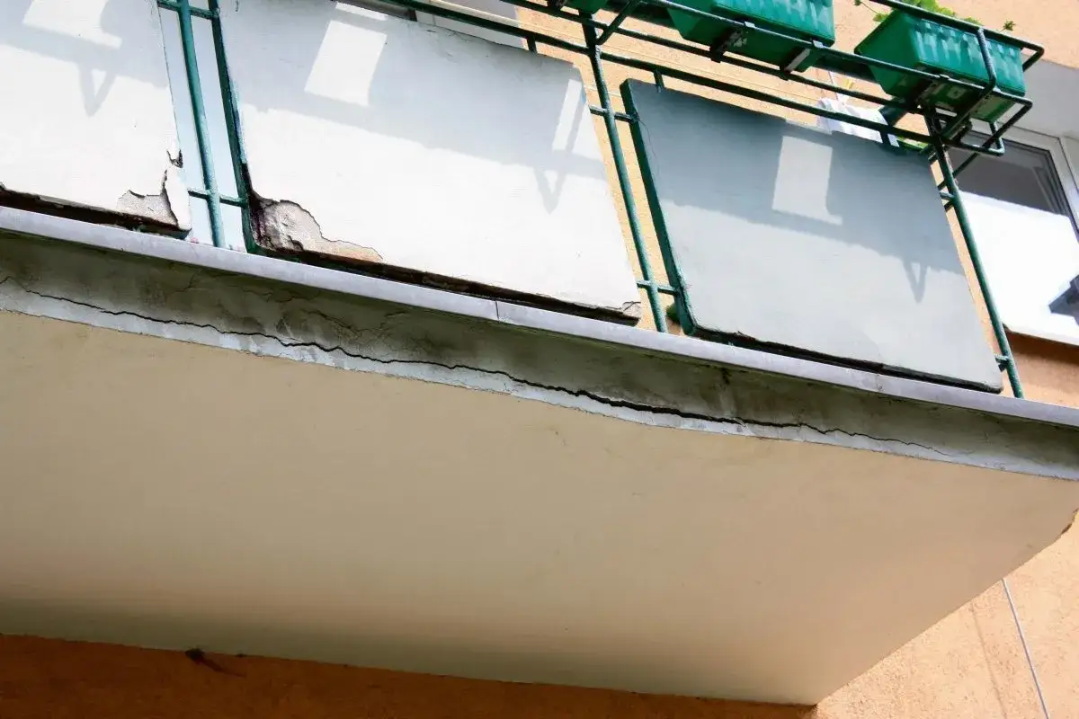 Zniszczony balkon w bloku z wielkiej płyty, z widocznymi pęknięciami i odpadającym tynkiem.