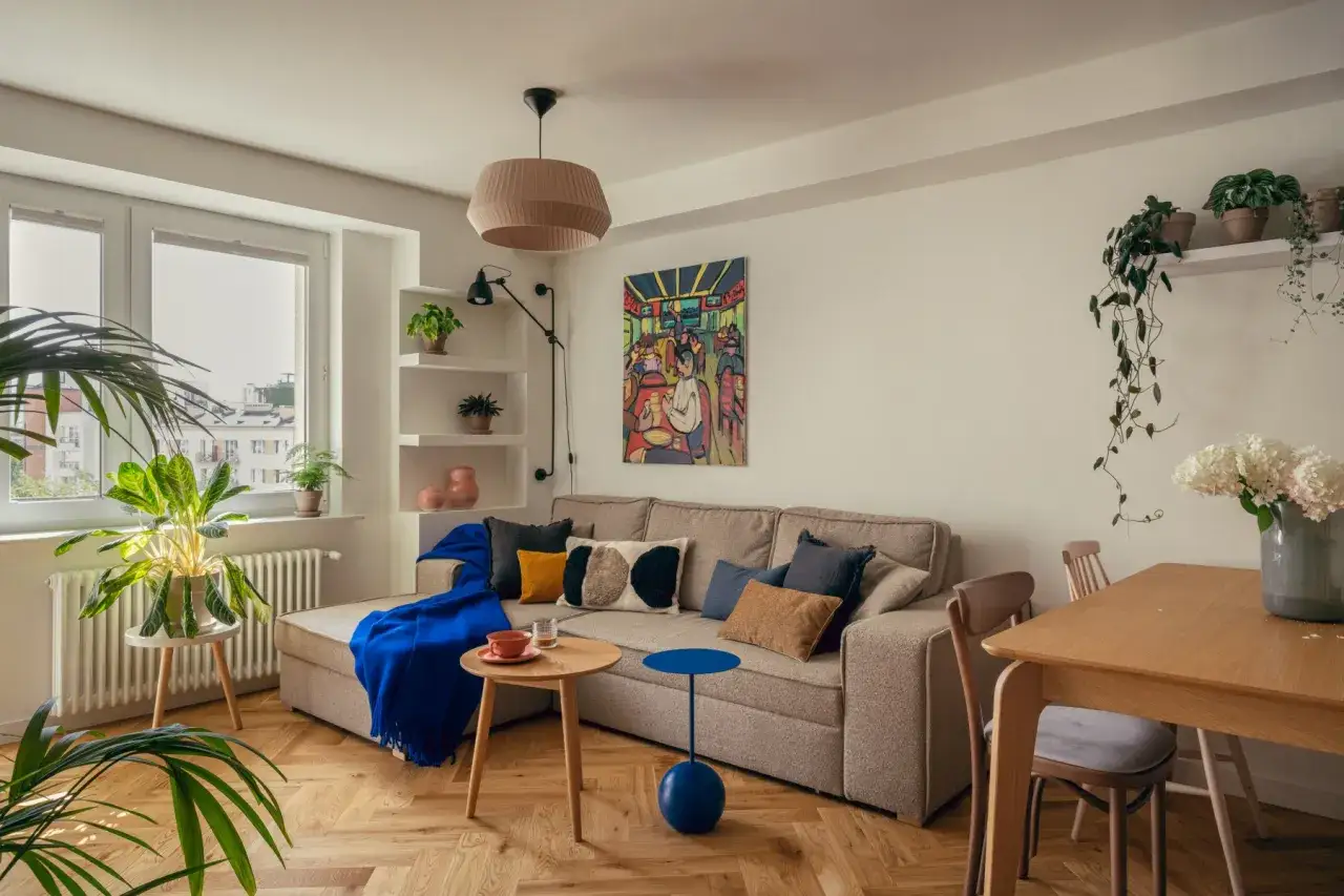 Przytulny salon z kanapą, stolikami i roślinami. Obraz na ścianie nawiązuje do klimatu mieszkań PRL.