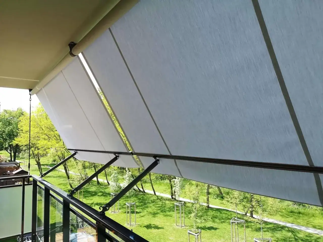 Zadaszenie balkonu w bloku bez wiercenia, chroniące przed słońcem. Rozłożony markiza z jasnego materiału, z widokiem na zielony park.