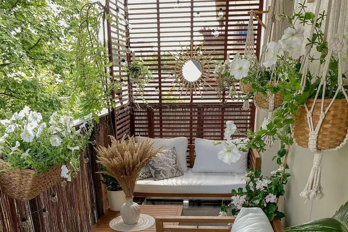 Przytulny balkon w bloku z drewnianymi zasłonami, sofą, białymi kwiatami i ozdobnym lustrem w kształcie słońca.